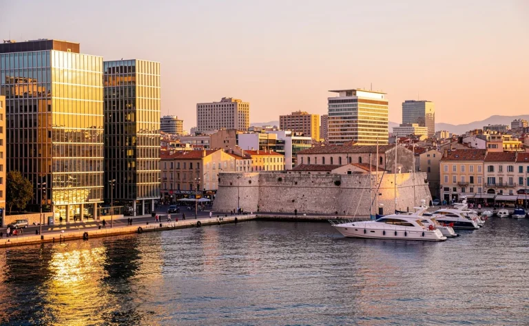 Panorama du port de Marseille au coucher du soleil, avec des immeubles modernes dominant le Vieux-Port et quelques bateaux amarrés.