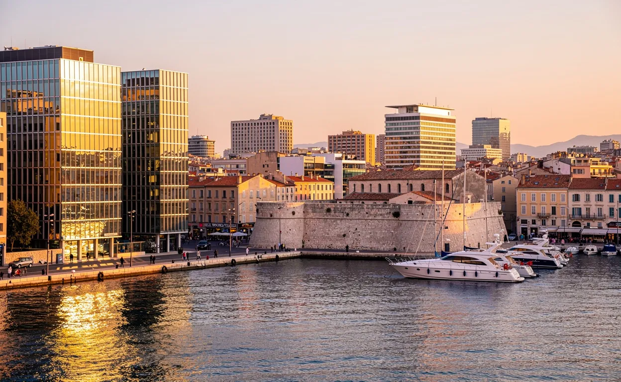 Panorama du port de Marseille au coucher du soleil, avec des immeubles modernes dominant le Vieux-Port et quelques bateaux amarrés.